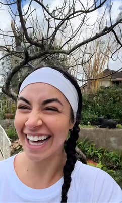 A woman with her hair tied up standing in front of a tree and smiling broadly.