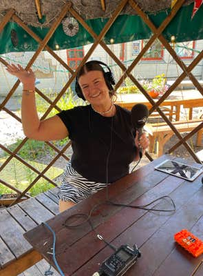 A woman sitting at an outdoor table posing for the camera with one arm raised, the other holding a microphone. She is wearing a headset.