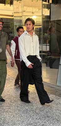 Actor Hudson Williams wearing a white decorated button up shirt and pinstripe pants, one hand in his pocket as he walks into a mall with the cast of I Love LA behind him.