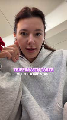 A woman with red hair and a grey hoodie talking at her camera, leaning down slightly. Text overlay reads, "Trippin with Tarte off the [sic] a bad start."