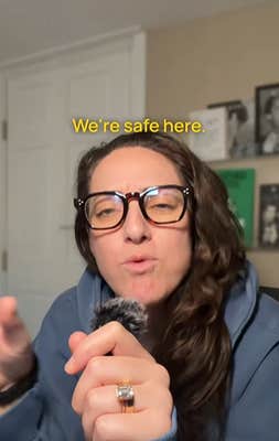 Woman with long brown hair and big glasses holding a lapel mic in her hand and talking to the camera in her home. Text overlay reads, "We're safe here."