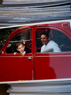 A small child and his father sitting in a bright red classic car, the little boy looking out the window while his dad laughs and watches him.