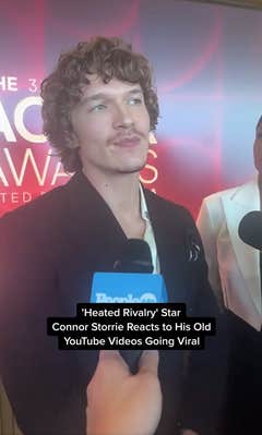 Connor Storrie on the red carpet at the Actor Awards nominations announcement. He is speaking into a mic. Text overlay reads, "Heated Rivalry star Connor Storrie reacts to his old YouTube videos going viral."