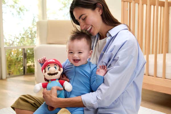 A smiling woman holding a baby in a plain blue onesie, holding a Mario plushie in her free hand. Her thumb appears to be bent backward in an uncomfortable way.